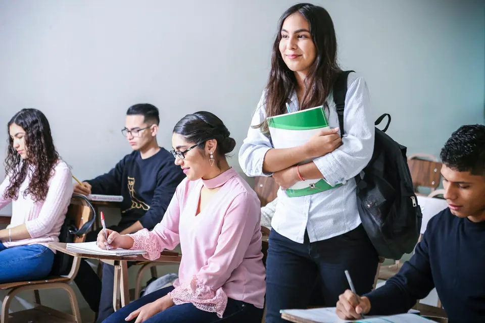 Estudante em pé numa sala de aula, com cadernos no braço, ao lado de colegas sentados